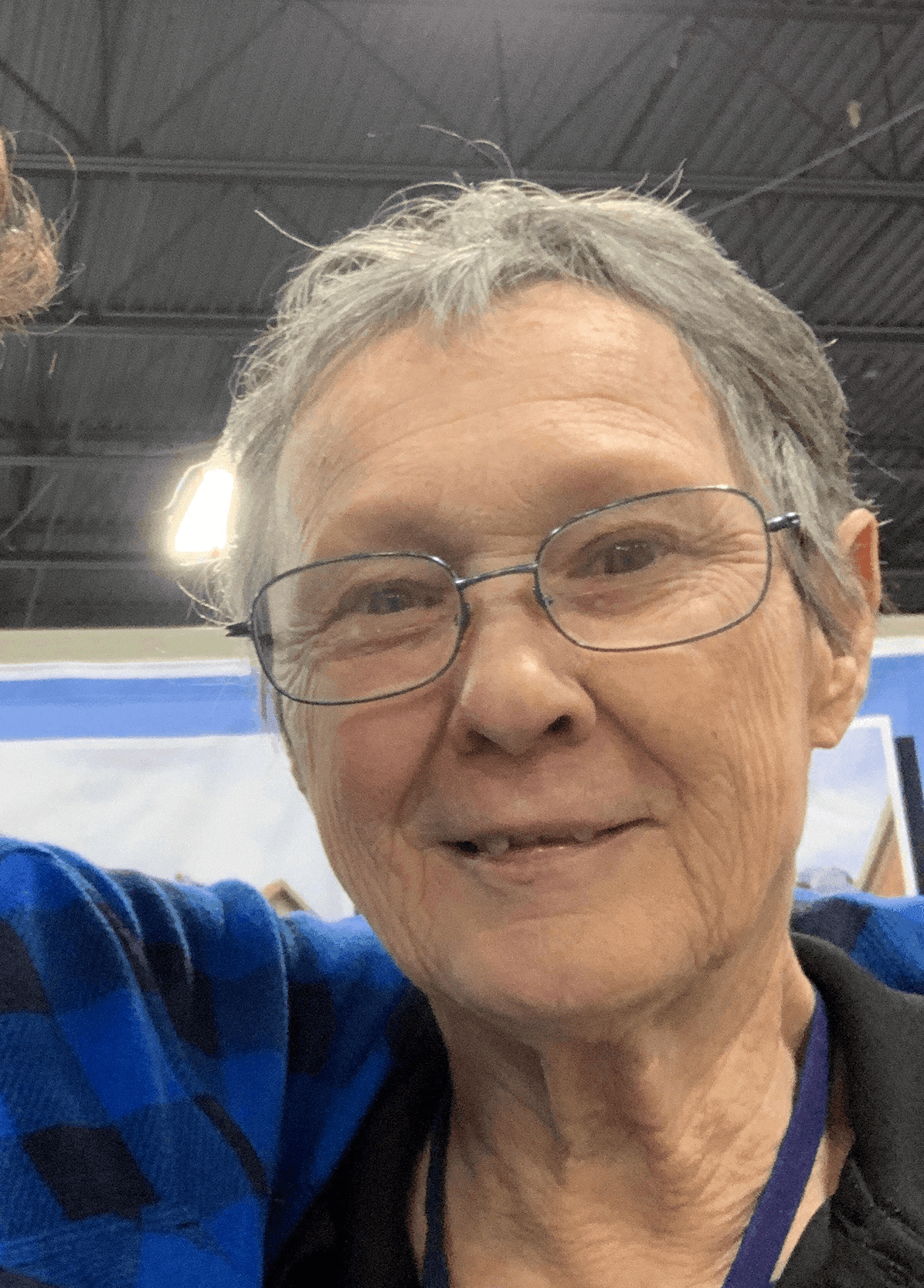 Older person with glasses smiling in an indoor setting, wearing a blue plaid shirt.